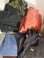 Top view of assorted clothing including black shirt with yellow Medusa logo, green Ralph Lauren Polo shirt, salmon colored pants, blue Levi's jeans with patch, black Lacoste polo shirt with logo