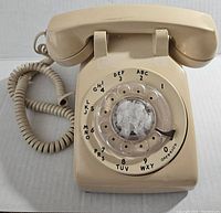 Front angled view of beige rotary dial phone showing handset, rotary dial with numbers and letters, and coiled cord.
