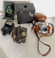 Five vintage camera-related items displayed on white surface: green folding camera, black box camera, brown leather camera with strap, small vintage camera, Minolta Auto 200X flash