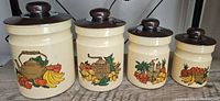 Set of four vintage Kromex stackable canisters arranged in descending size order, showing rusty cream color and lids with matching dark brown knobs and colorful fruit and teapot graphics on the side.