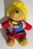 Front view of the vintage Paddington Bear plush toy dressed in yellow hat, red shirt and denim overall with Craftsman logo, new with original tags.