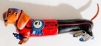 Side profile photo showing the elongated dachshund figurine painted in red and black racing attire with a blue #1 ribbon that reads 'Winner of the race!' on its side.