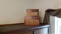 Two decorative wooden boxes stacked on a piece of furniture against a cream wall. Both have latch closures and wood laminate front panels.