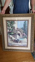 Framed art print depicting a Tuscan villa courtyard with stone fountain, flowers, and stairway, held upright to show entire piece