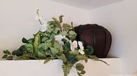 Photo of large dark brown woven wood pot with lid, placed on high ledge near green foliage and white flowers.