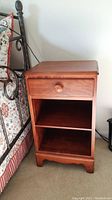 Front view of the wooden night stand showing drawer and shelves, placed beside a bed.