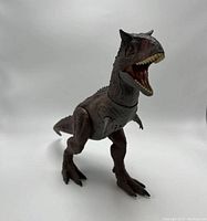Front angled view of articulated dark brown Carnotaurus Toro figure from Jurassic World Camp Cretaceous series showing open mouth and front limbs.