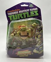 Front view of the Michelangelo Teenage Mutant Ninja Turtles action figure in original packaging, showing the figure holding nunchuks inside a clear blister pack with purple cardboard backing and TMNT branding.