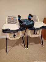 Two white plastic baby high chairs with attached trays and teddy bear pattern backrests, positioned side by side with a black and blue booster seat in front.