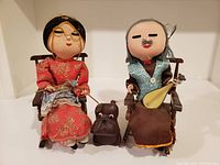 Pair of stuffed Chinese husband and wife dolls seated in wooden chairs with small stuffed puppy dog between them, showing overall condition and scene