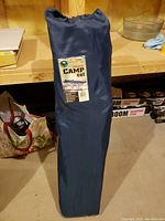 Blue carry sleeve holding the folded camp cot. Sleeve is cylindrical with a drawstring top and a label attached.
