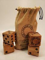 Six oversized wooden dice with black pips and one die showing 'Snake Eyes Yard Dice' logo face, arranged in front of a drawstring burlap carry bag.