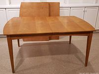 Front view of rectangular solid wood dining table with extension leaves leaning behind it. Table surface shows marks and scuffs.