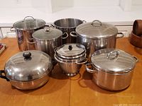 All items arranged on a table showing large stainless steel pots with lids, a colander nested in a steamer insert, and a covered pan with black handles and knob. Items show signs of use and surface marks.