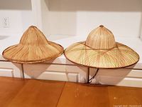 Photo showing two traditional Chinese bamboo hats side by side, one plain bamboo and one with red and green woven pattern on brim and dome, both conical shape with wide brims.