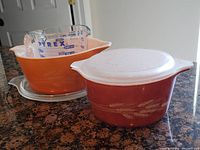 Photo of two vintage Pyrex bowls, orange and maroon, with maroon one having a white plastic lid, and a clear Pyrex glass measuring cup with blue markings inside the orange bowl.