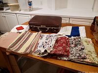 Brown hard shell suitcase on table with various folded fabrics laid out in front of it.