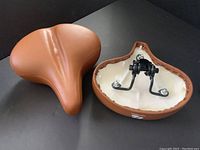 Two views of one brown bicycle saddle seat showing the top smooth leather surface and bottom plastic shell with metal mounting hardware.
