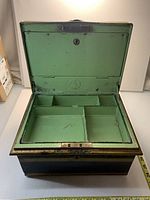 Front view of antique metal lock box with lid open showing green interior and multiple compartments.