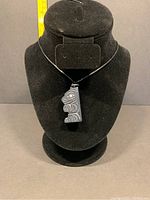Full frontal view of the black argillite bear pendant hanging on a black cord displayed on a black bust stand with measuring tape in background showing size.