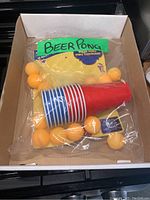 Photo of the complete Beer Pong game set including stacked red and blue disposable cups and orange ping pong balls inside sealed plastic packaging, placed in a cardboard box.