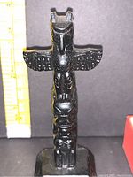 Front view of the 4 inch black carved totem pole showing detailed bird wings and stacked faces, standing on a black base with original box partially visible.