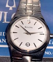 Close-up of the Bulova watch face showing white dial, silver hour markers and hands, date window at 6 o'clock, and Bulova logo.