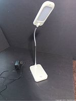 White LED desk lamp shown standing upright with flexible neck extended, power adapter plugged in and placed beside it.