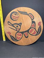 Front view of circular carved wood panel with wolf and moon motif, scale shown by ruler at side.