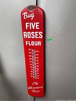 Full view of red metal wall-mounted thermometer with white “Buy Five Roses Flour” lettering and central glass tube