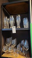 Top shelf displays four water glasses with vertical cuts and six controlled bubble beer glasses with thick clear bases containing internal bubbles, bottom shelf shows four Riedel wine glasses with rounded bowls and stems, and two wide Hennessy cognac glasses with short stems and rounded bowls.