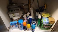Under-sink cabinet with various cleaning supplies arranged inside including wipes, sprays, creams, and sponges.