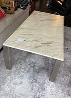 Rectangular coffee table with white marble top showing gray veining, placed on a carpeted floor next to a sofa and chair, steel legs are visible in brushed finish.