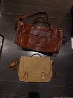 Two bags laid flat on dark floor: brown leather duffle bag with front pocket and beige Frank and Oak canvas messenger bag with leather straps