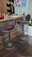 Wide view showing the pair of matching adjustable bar stools in front of a counter, with black nylon velvet cushions and dark metal powder coated bases.