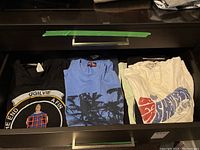 Drawer with nine graphic t-shirts folded in three rows, showing various designs and colors