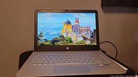 Front view of open HP ENVY laptop with screen on showing scenic wallpaper, silver keyboard, power cable connected