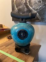 Frontal view of pot showing turquoise crackle glaze, circular emblem, black handle and pedestal