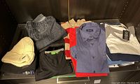 Overview of men's clothing including folded shirts, sweaters, knit toques, and black denim trousers on display.