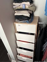 Stack of folded men's clothes on top of a tall narrow drawer unit, showing multiple folded pieces from shirts to jeans.