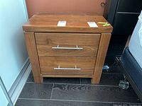 Front view of walnut finish nightstand with two drawers and silver metal bar handles on dark floor.