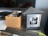 Front and side view of metal outdoor lantern with window cutouts and a cardboard box behind it.