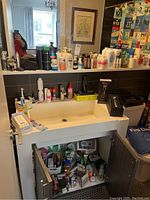 Wide view of bathroom sink and counter showing assortment of personal care products, lotions, sprays, and first aid items stored on the sink counter and under the cabinet.