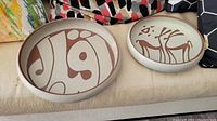 Two artisan ceramic bowls side by side, one larger with abstract calligraphy design, one smaller with deer motif