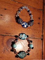 Two handcrafted expandable bracelets on wooden surface; one with large textured blue and clear art glass beads, and the other with turquoise patterned glass beads and smaller black and brown beads.