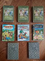 Seven vintage Bobbsey Twins hardcover children's books arranged on wooden floor, showing front covers and back covers with varying degrees of wear and age.