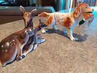The set of two vintage ceramic figurines showing a doe and fawn figurine alongside a dog figurine holding a duck in its mouth, placed on a granite countertop.