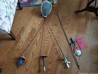 Full spread of vintage fencing equipment including three foils, fencing mask, additional blade, glove, and booklet on hardwood floor.