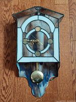 Front view of the clock showing stained glass face and brass pendulum on wooden floor.