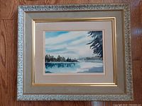 Frontal view of vintage watercolour painting showing a harbor scene, water, tree silhouettes, blue sky with clouds. The painting is matted and surrounded by a decorative carved wood frame with gold and beige tones.